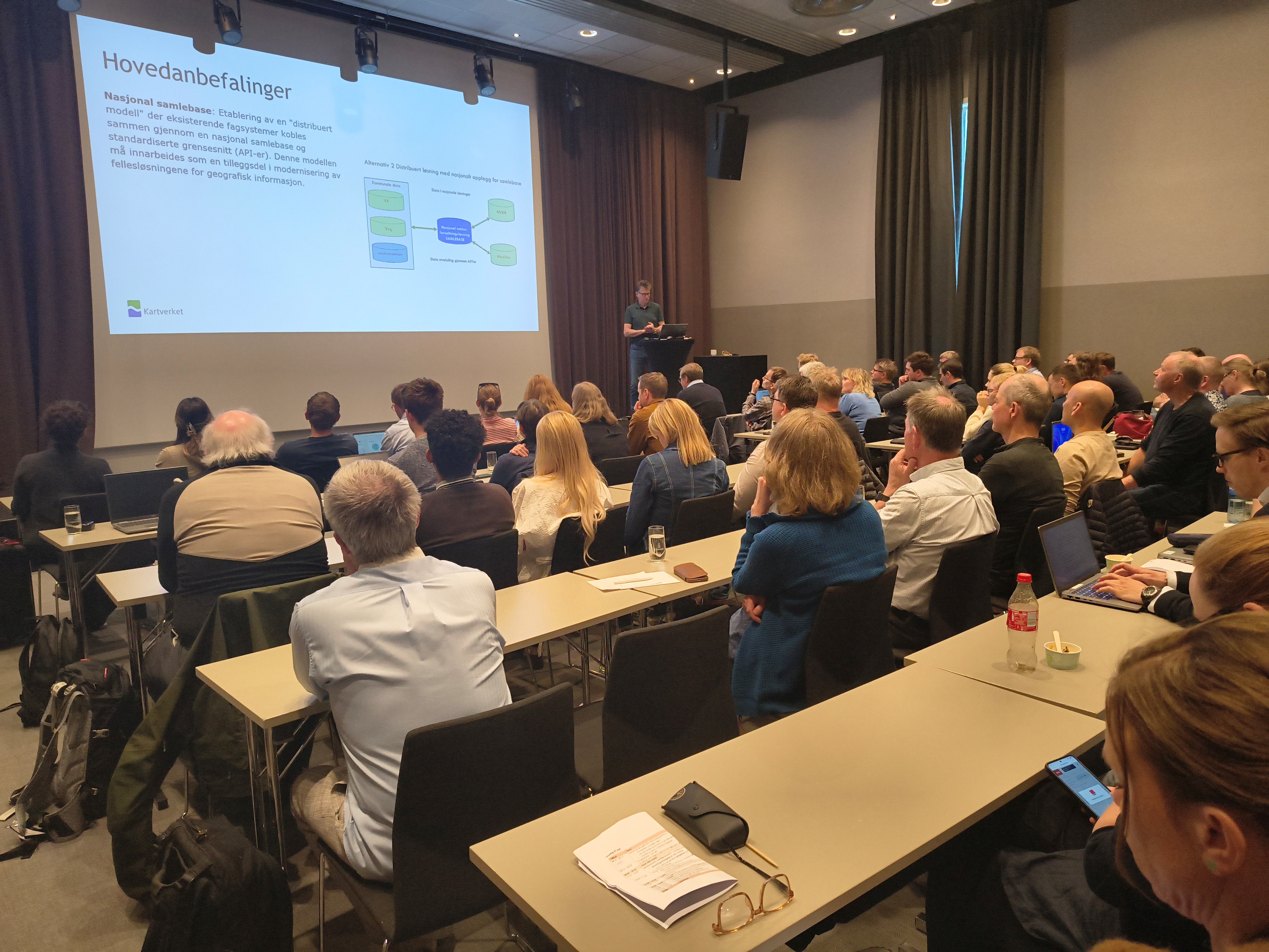 Mann st&aring;r p&aring; en scene foran en fsittende forsamling og presenterer noe p&aring; en skjerm.
