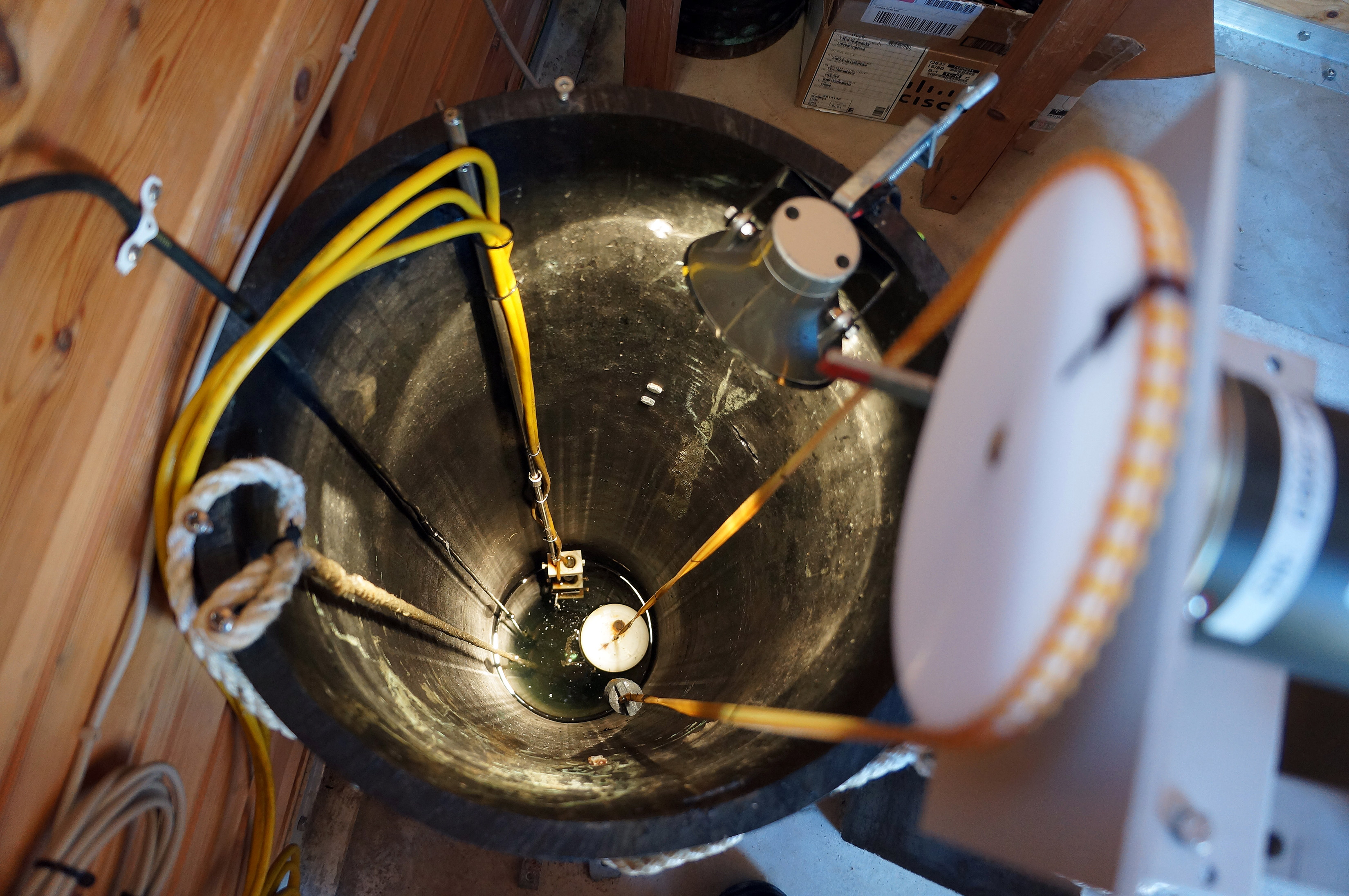 Flottørmåler, et rør med en trang åpning i bunnen er plassert ned i sjøen. Over røret henger et tannhjul med et kjede som har en flottør i den ene enden og en motvekt, et lodd, i den andre. Foto.