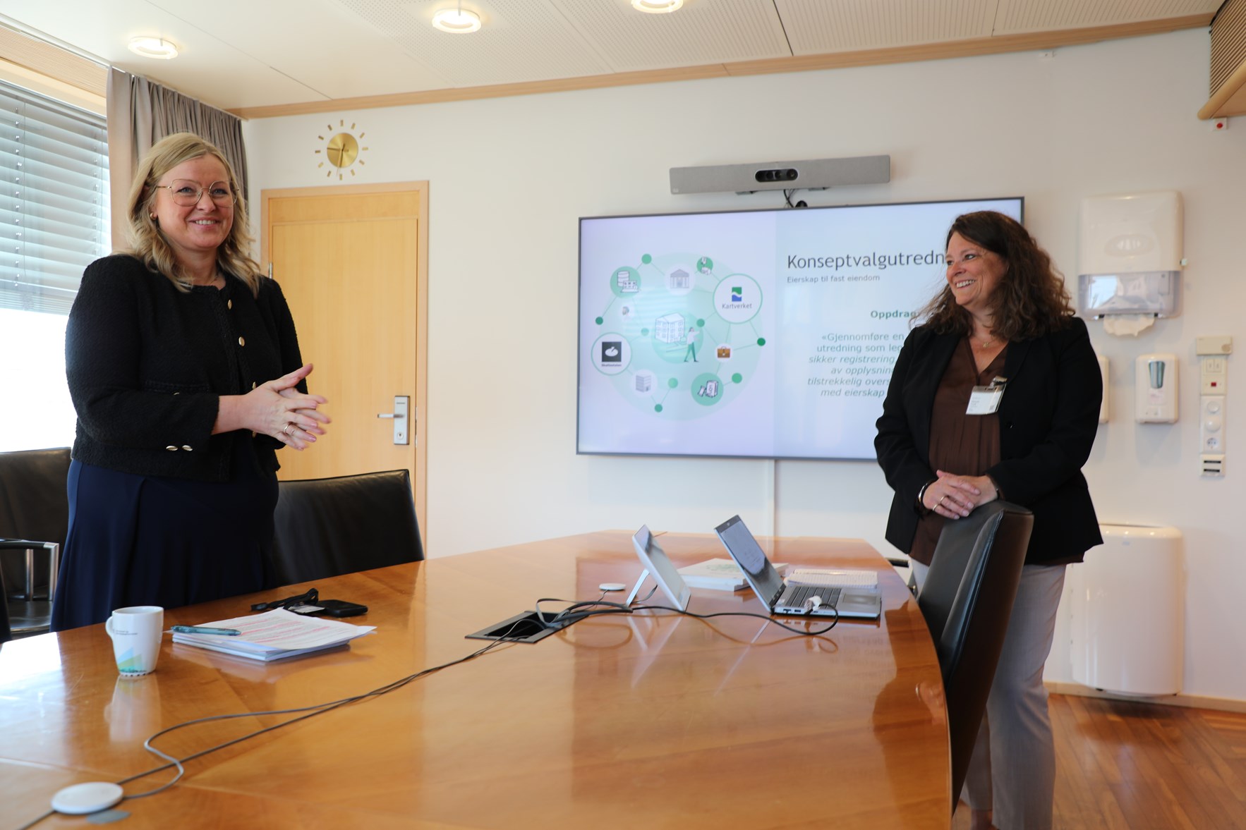 En kvinnelig statsråd og en kvinnelig divisjonsdirektør i Kartverket på hver sin side av et bord der en storskjerm ved enden av bordet viser en powerpoint om Kartverkets konseptvalgutredning (Foto: Synne Storvik/ Kartverket)