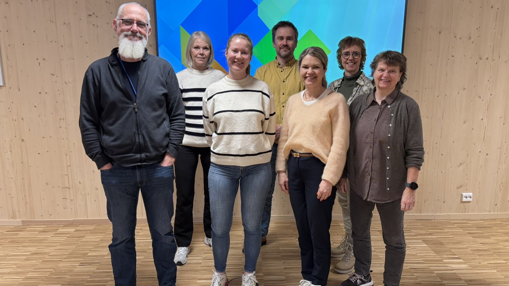 To menn og fem kvinner står foran en innendørs vegg