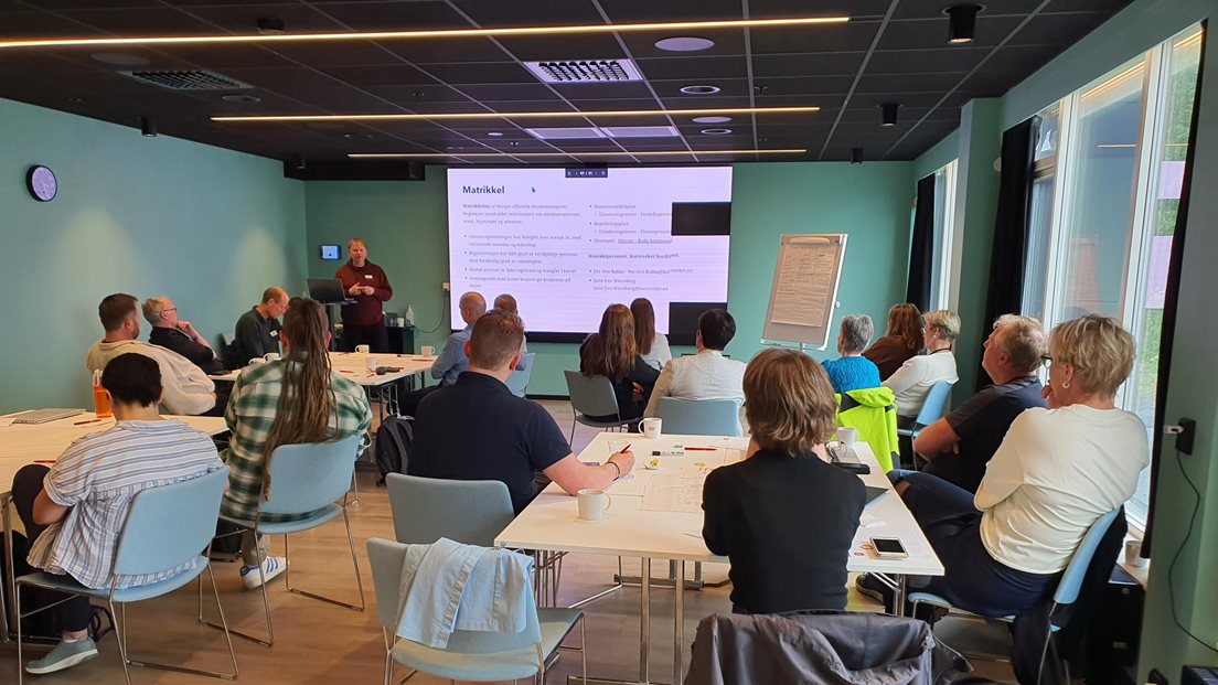 Mange personer sitter i et rom og ser på foredragsholder og torskjerm