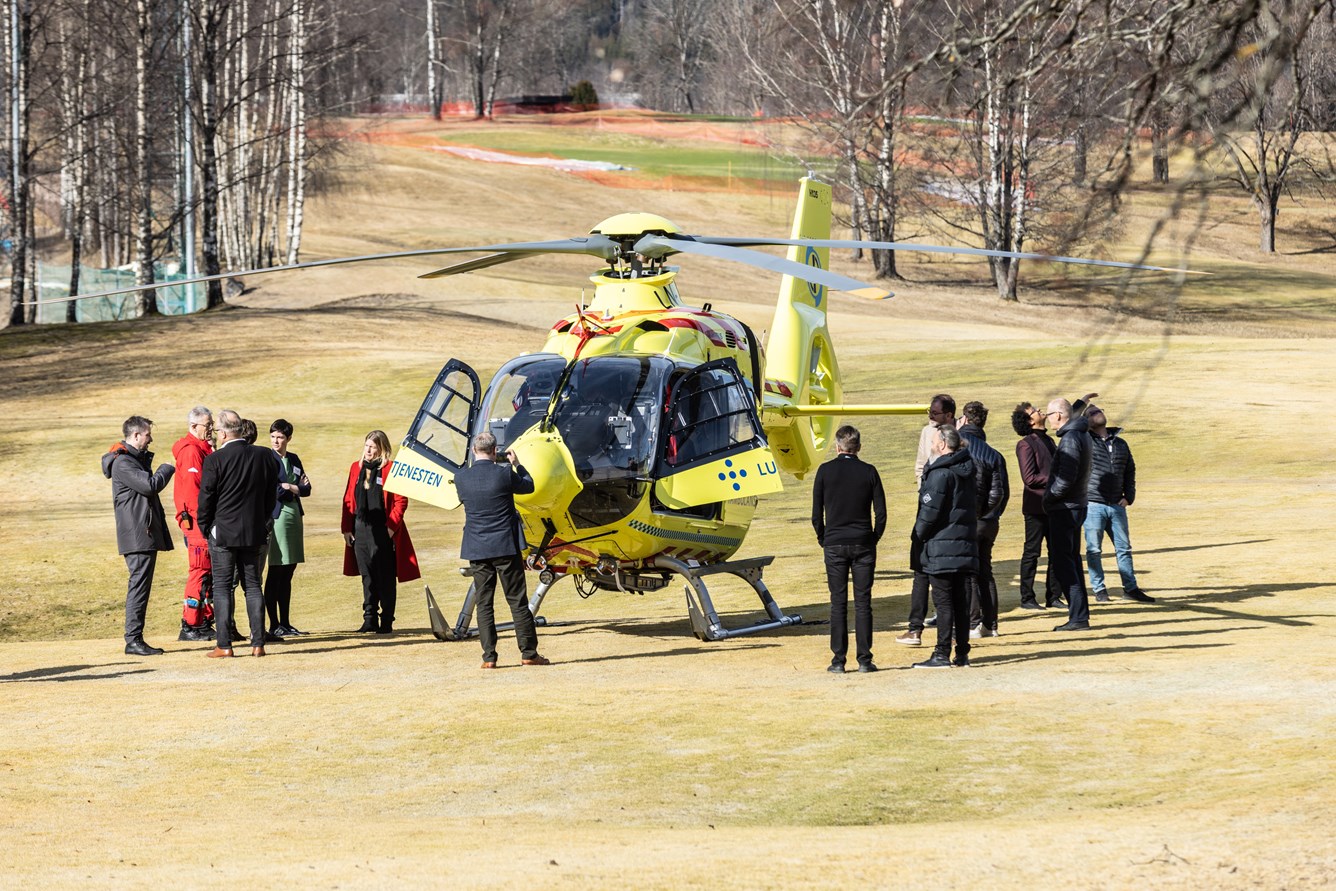 En gruppe mennesker står rundt et gult helikopter fra Norsk Luftambulanse som står på en gressplen.