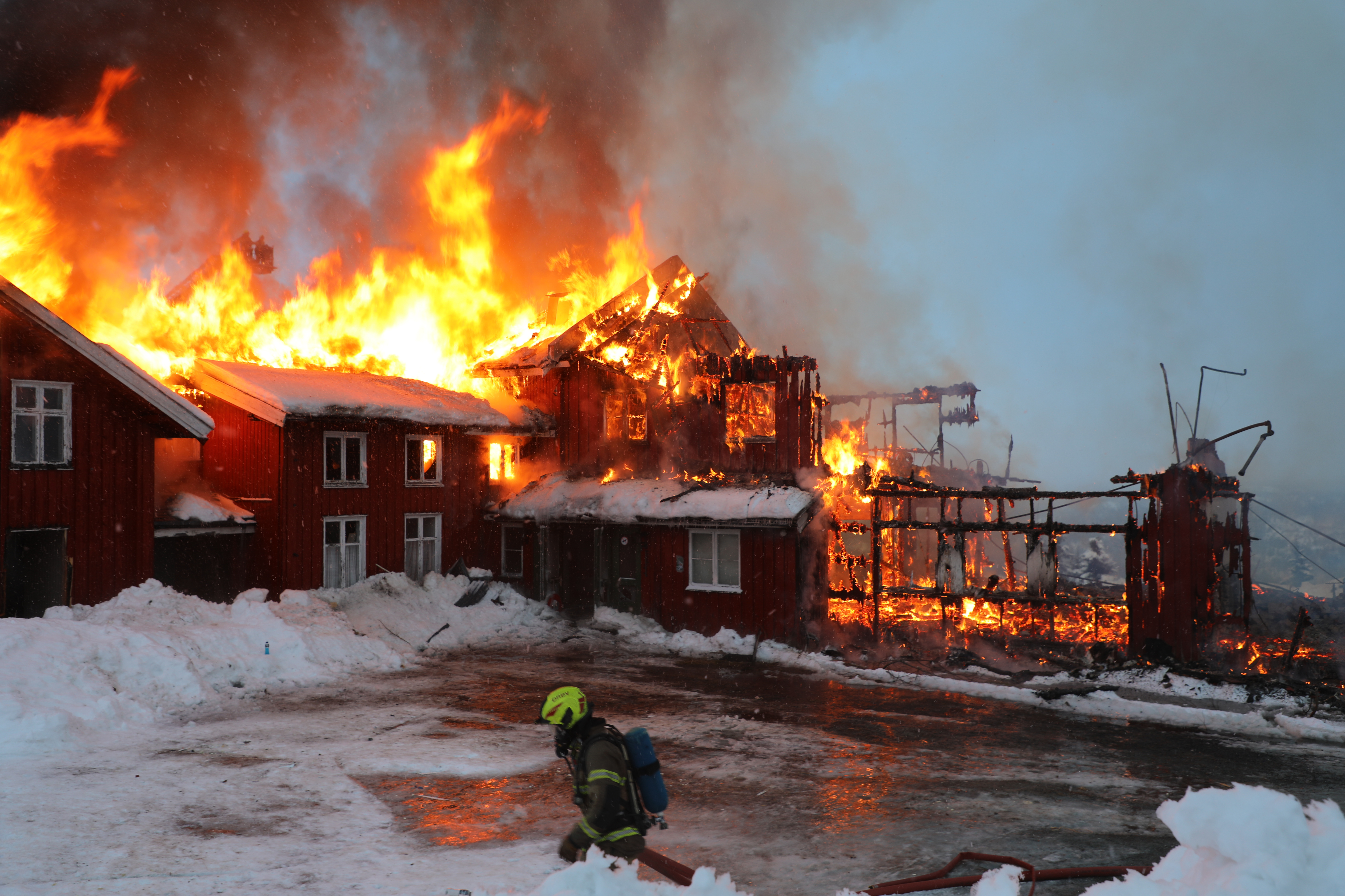 En brannmann trekker en slange over en islagt plass, foran et stort hytteanlegg som blir brent ned i en kontrollert nedbrenning på Norefjell. Det står flammer opp av taket i bakgrunnen.