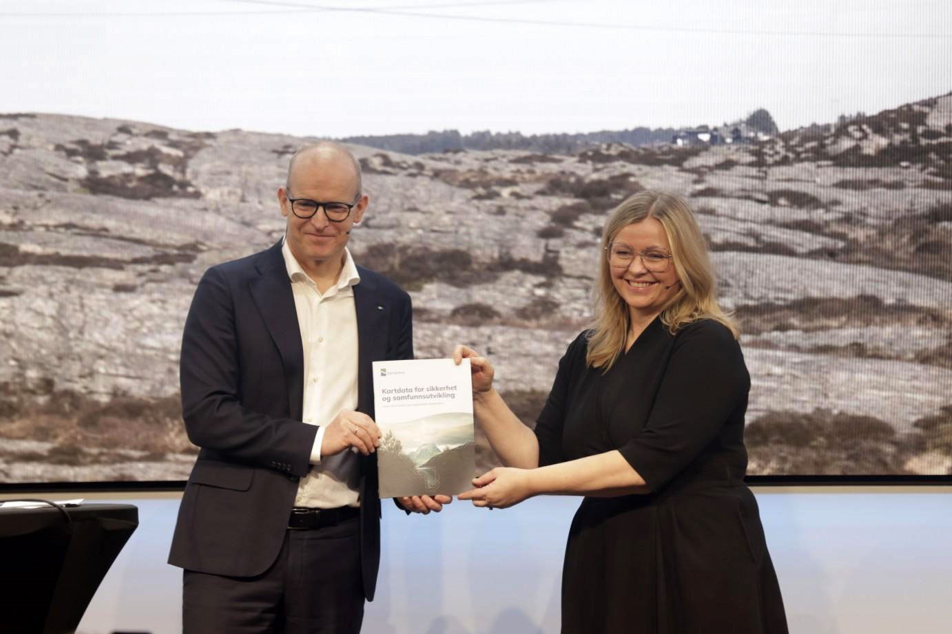 Kvinnelig statsråd mottar Kartverkets statusrapport fra mannlig kartverkssjef på et podium