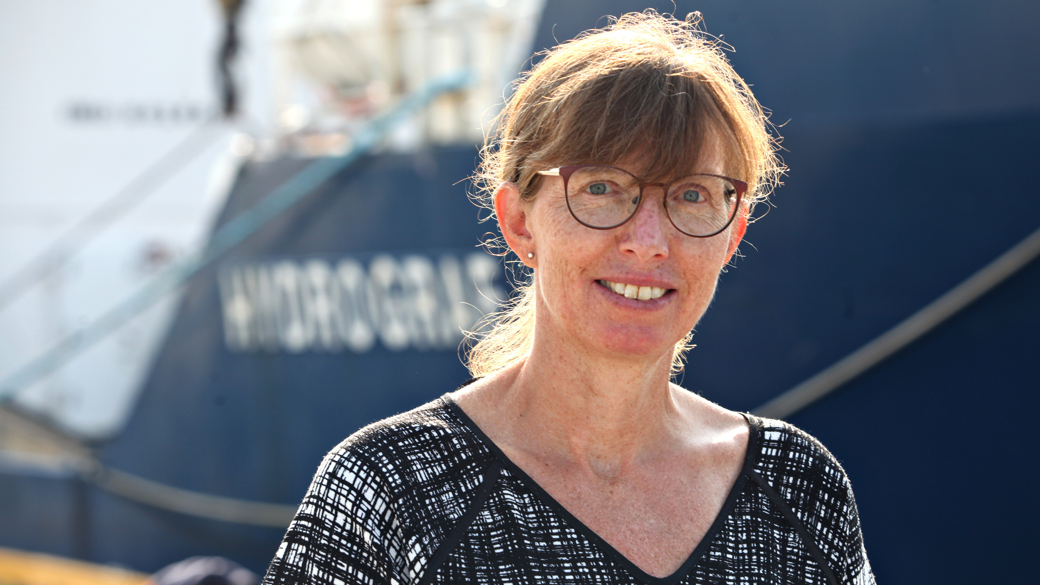 Dame med brunt h&aring;r og briller st&aring;r foran et st&oslash;rre fart&oslash;y i ei havn. Det er sol ute. Foto.