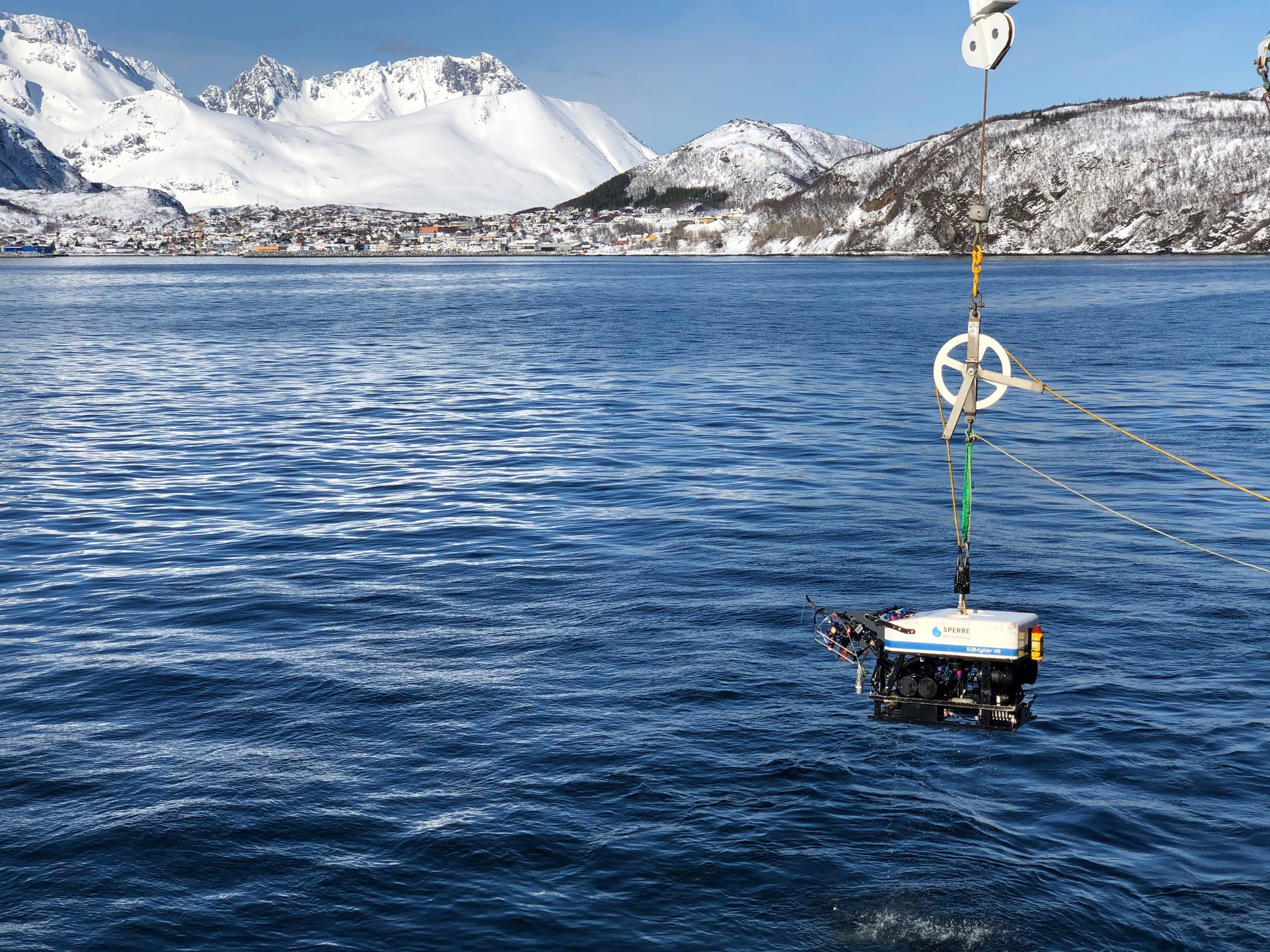 En undervannsfarkost henger over vannflaten og skal senkes ned til havbunnen. Utsikt til snøkledte fjell og blå himmel over bebyggelse på Skjervøy.