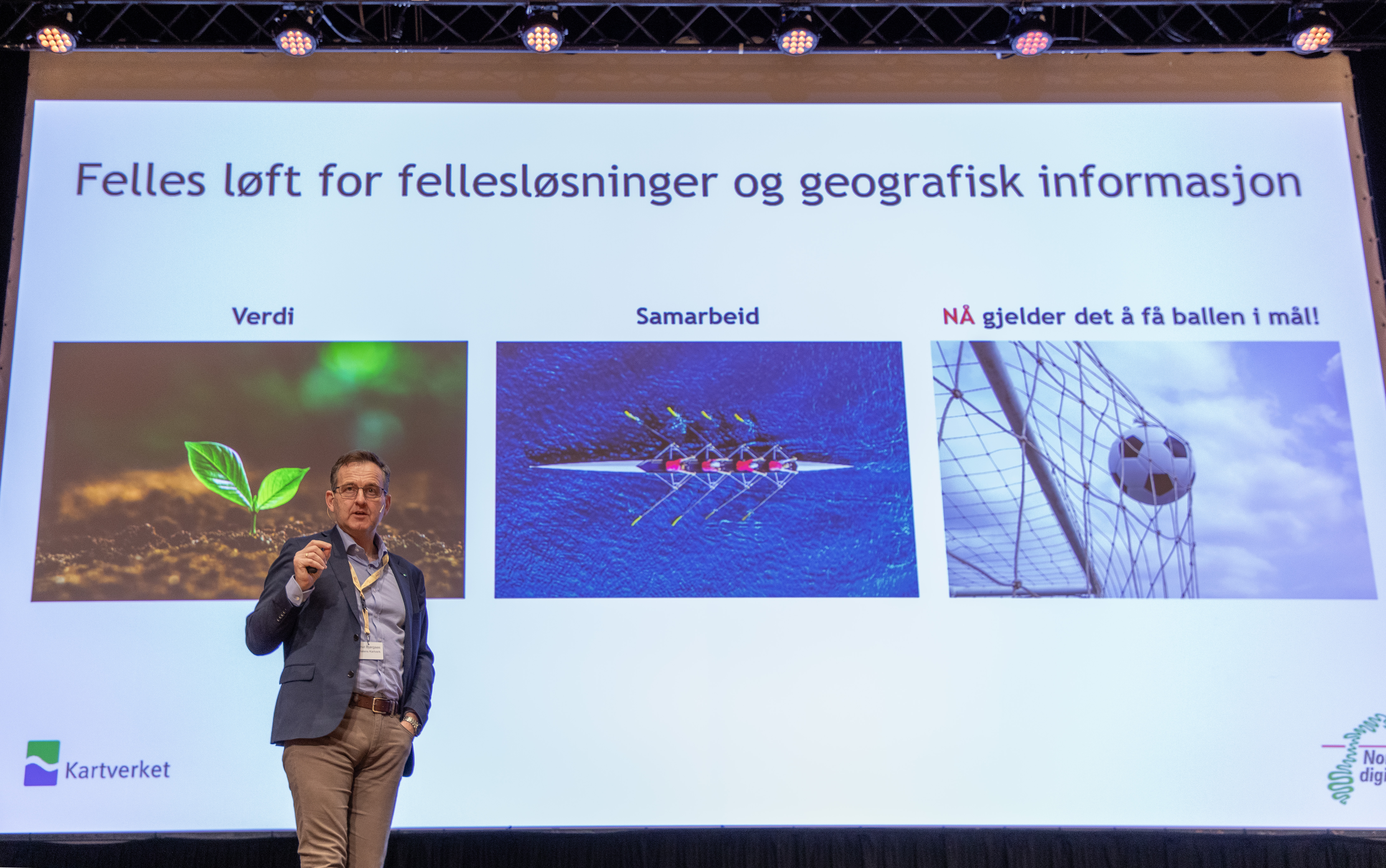 En mann st&aring;r p&aring; en scene foran en storskjerm og snakker engasjert. Skjermen viser en plansje med logo for Kartverket og Norge digitalt.