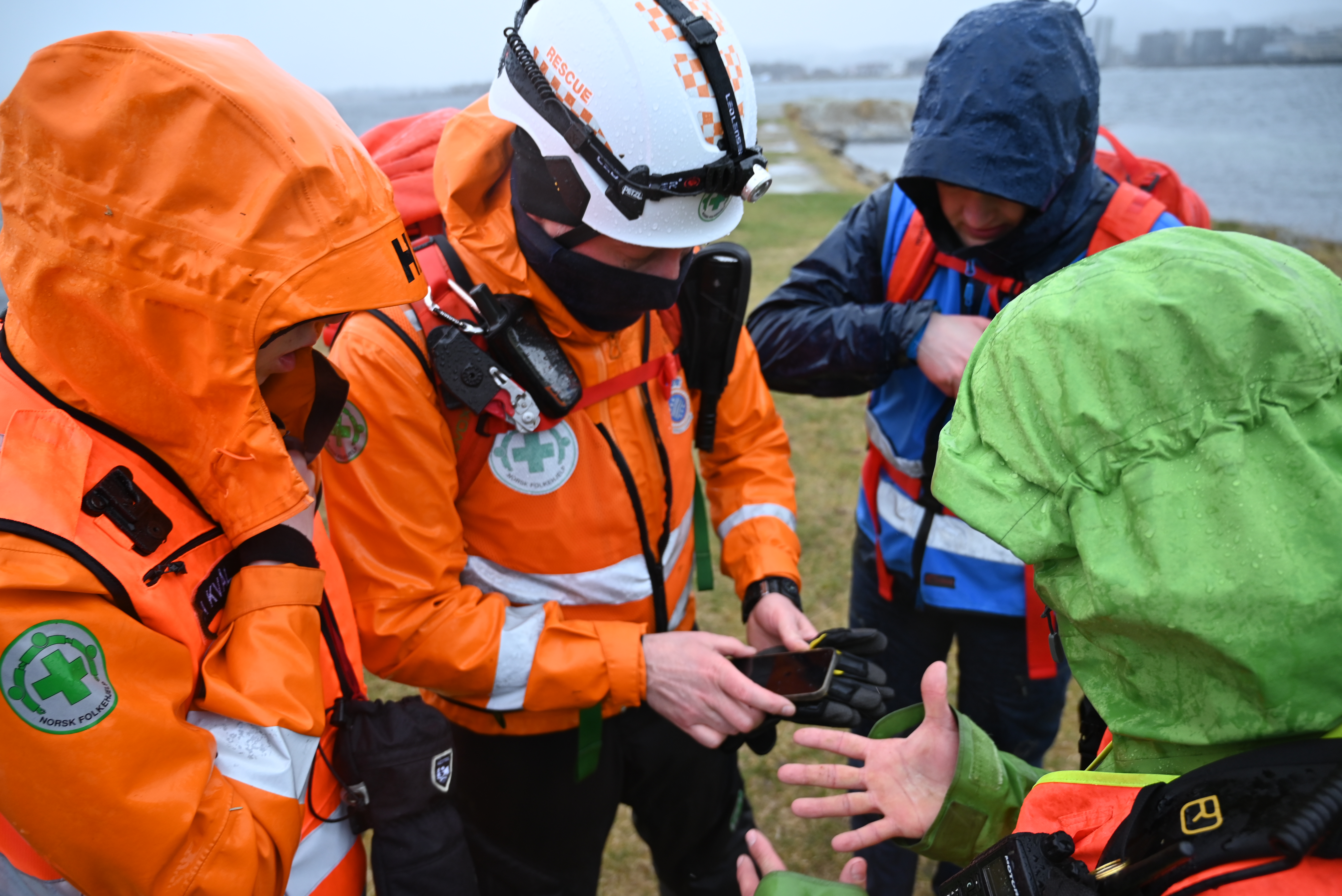 Redningsmannskap i oransje dresser med Norsk Folkehjelp-logo snakker med turister i dårlig vær.