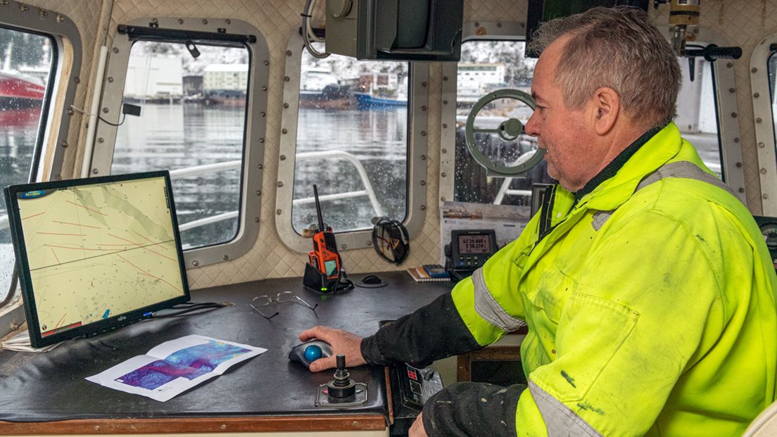 Mannlig krepsefisker iført gul jakke med refleks ser på kart på en skjerm om bord i fiskebåten sin. Foto.