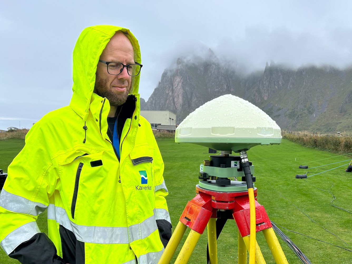 En person i gul jakke st&aring;r ute ved en GPS-stasjon i et gr&oslash;nt landskap, med fjell i bakgrunnen.