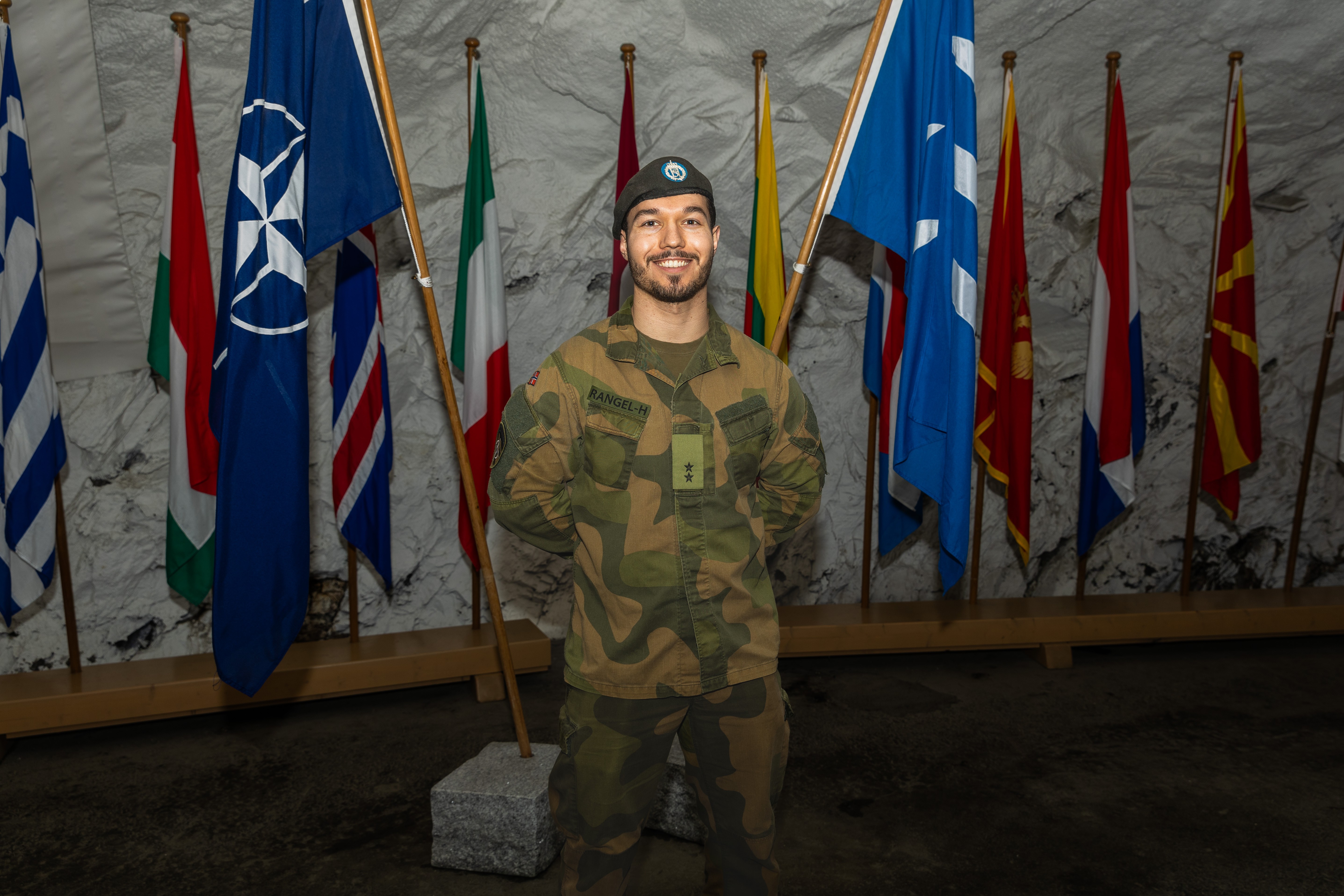 Mannlig soldat i uniform står mellom to flagg og smiler.