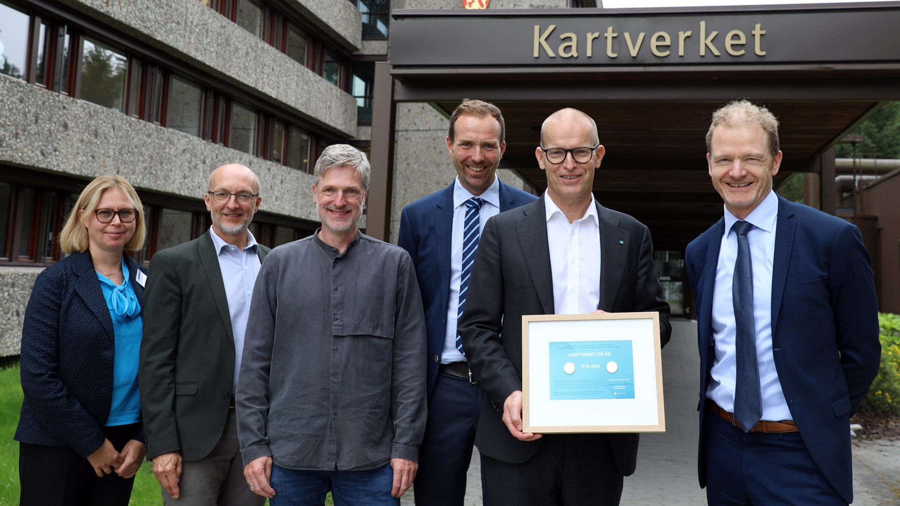 Representanter fra Norges Bank, kunstner Torgeir Husevaag, kartverkssjef Johnny Welle og visesentralbanksjef Pål Longva utenfor Kartverket på Hønefoss. Kartverkssjefen holder innrammet minnemynt.