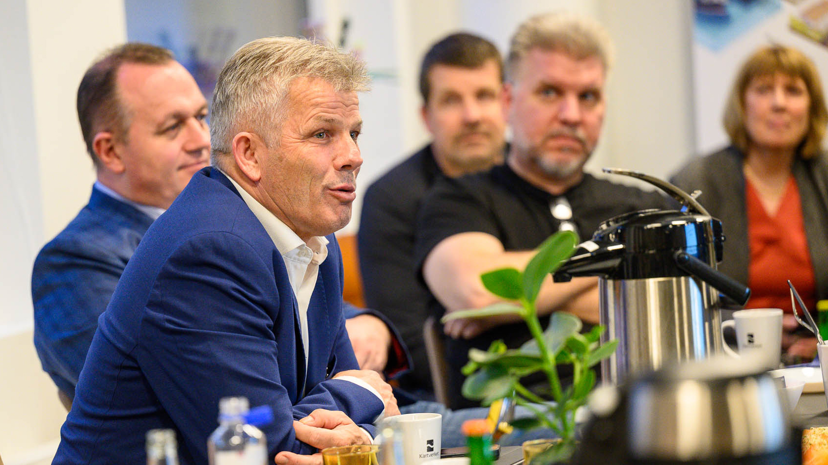 En mann i dress sitter ved et bord og snakker, med tre menn og ei dame uskarpt i bakgrunnen.