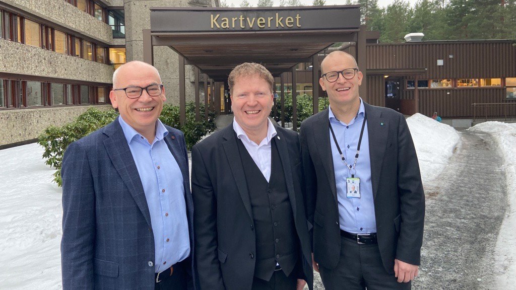 Tre smilende menn i dress og blå skjorte, Terje Dahlen, Sigbjørn Gjelsvik og Johnny Welle, står oppstilt utenfor hovedinngangen til Kartverkets hovedkontor på Hønefoss. Det er snø på bakken.