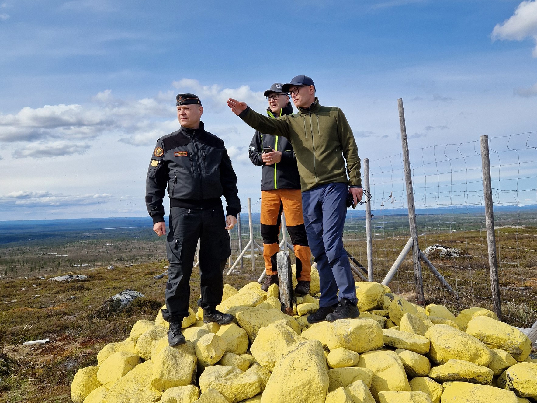 Tre menn står oppå ei gulmalt riksrøys hvor den ene peker utover landskapet.