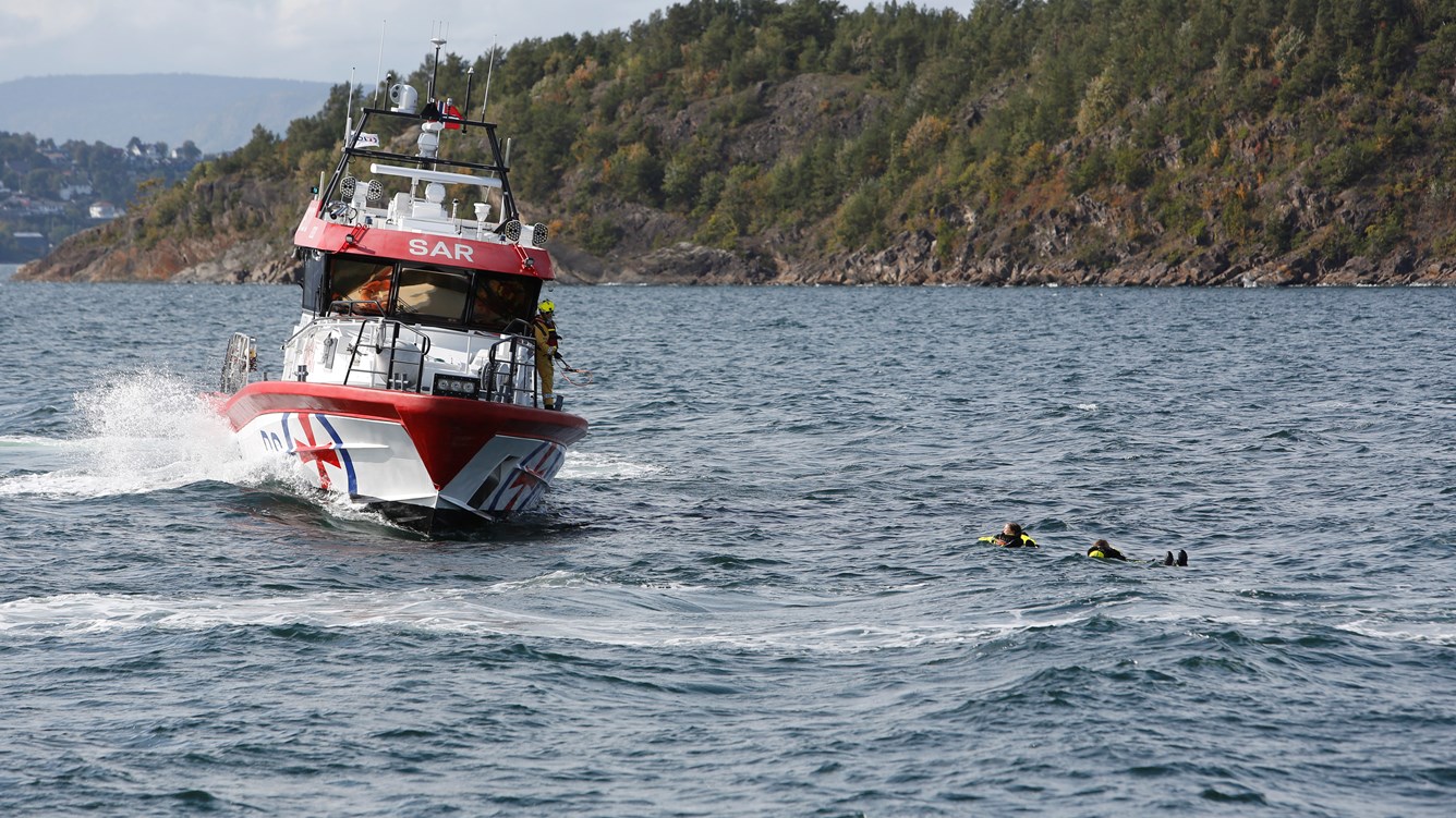 Redningsskøyte på vei for å redde to personer i flytedrakt som ligger i vannet.