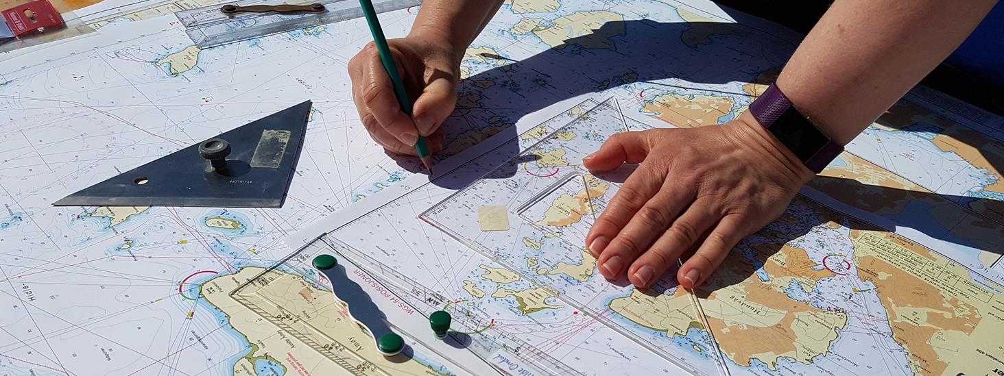 Et skolekart (sjøkart) ligger oppå et hovedseriekart. To hender plotter inn kurs med blyant og linjal. Foto.