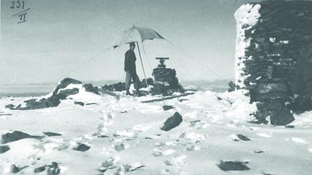 Fjellvarde og landmåler med teodolitt på målebordet som beskyttes av parasoll. 