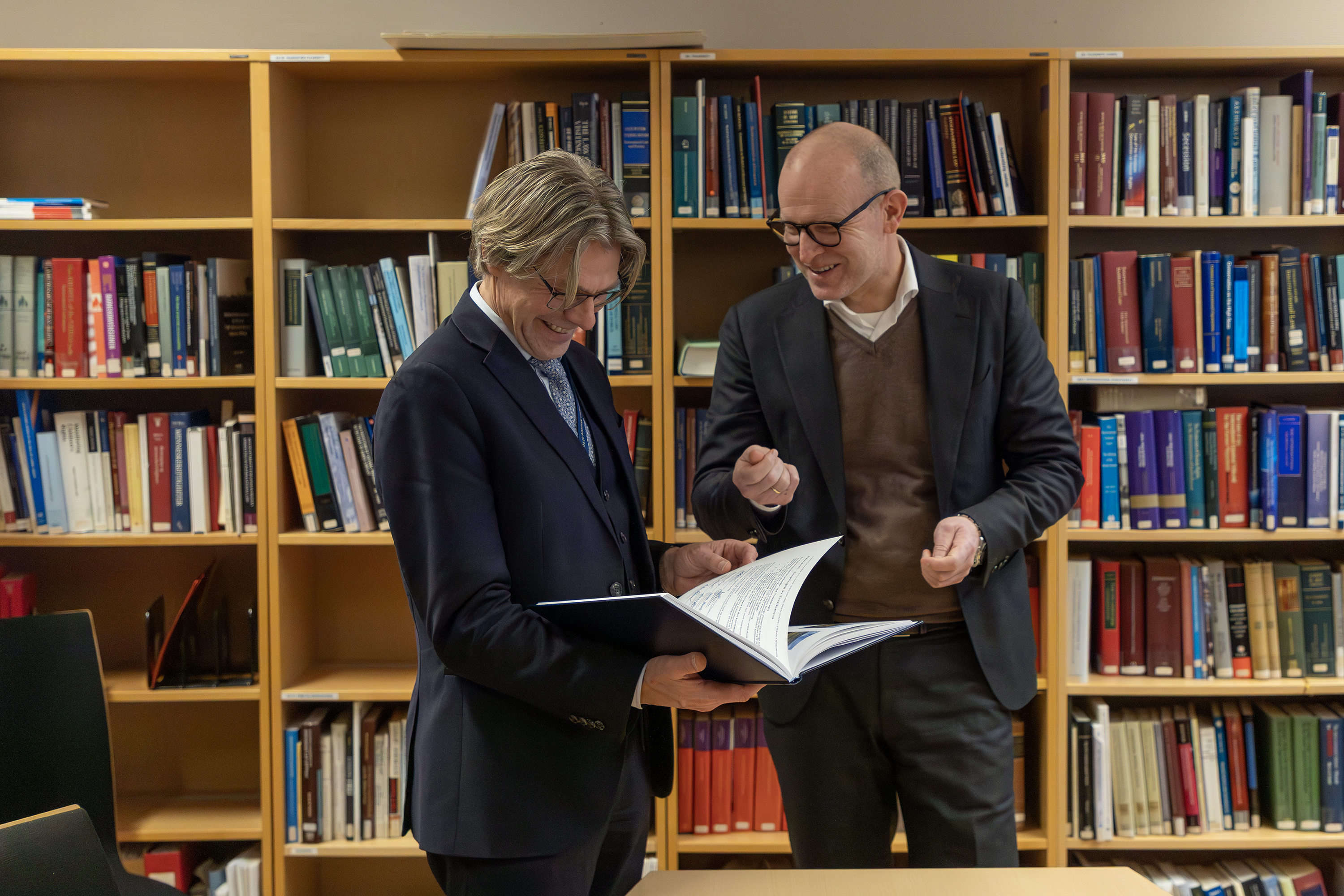 To menn i dress står med en perm foran bokhyller.